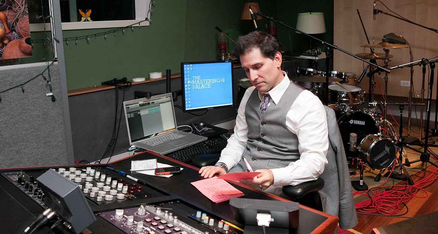 Dave Kutch sat in a suit in a recording studio, with a drum set behind him and a pen and paper with notes on that he is looking at Dave Kutch sat in a suit in a recording studio, with a drum set behind him and a pen and paper with notes on that he is looking at
